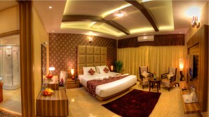 A hotel room with a large bed, a sitting area with chairs, a desk with a lamp, a shower, and a large window with curtains. - Jal Mahal Resort & Spa, Mysore (Mysore)