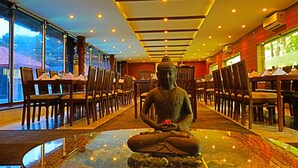 A statue of a seated figure in a restaurant with tables and chairs arranged around it, large windows on one side, and a ceiling with recessed lighting. - Jal Mahal Resort & Spa, Mysore (Mysore)
