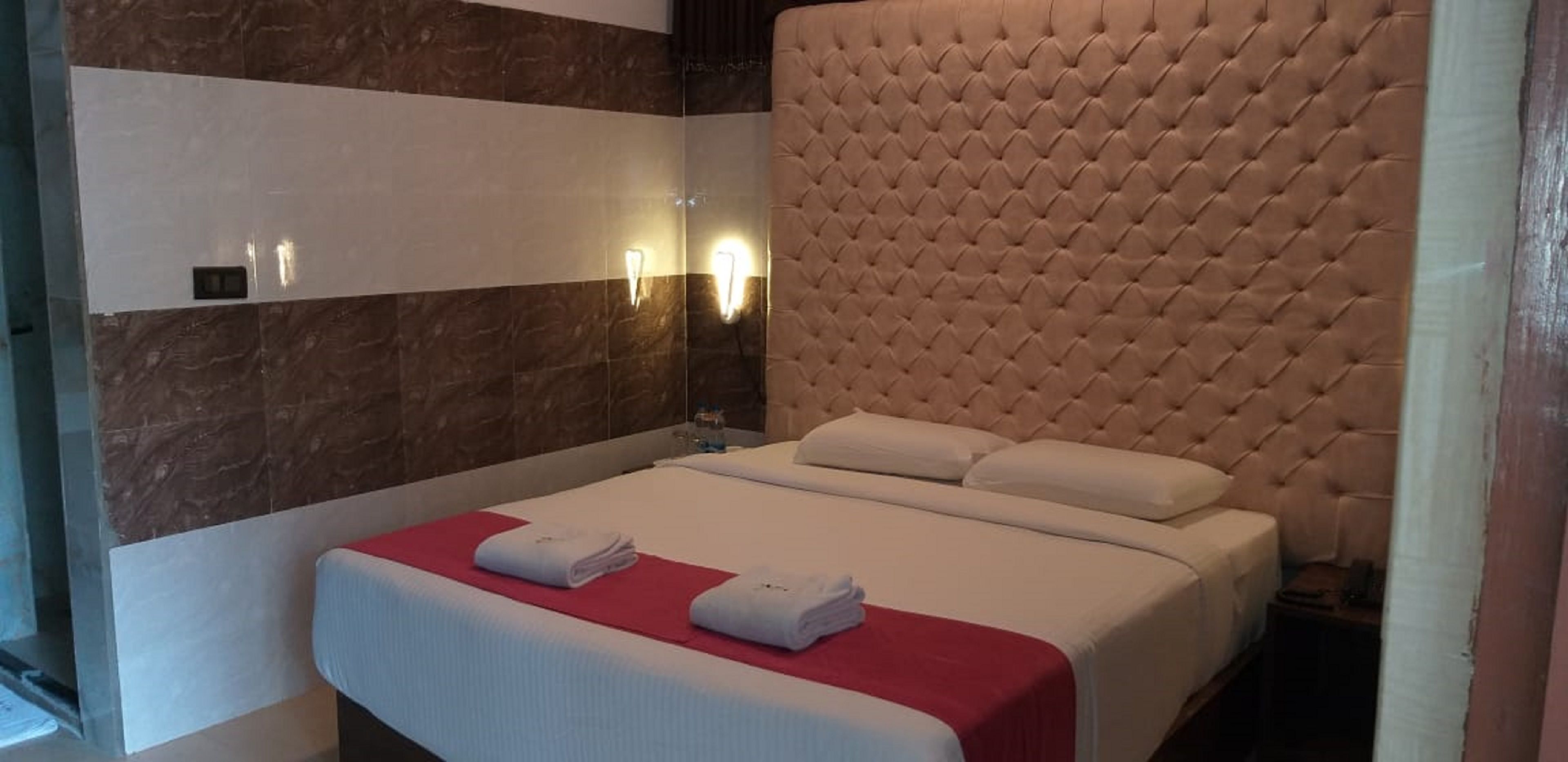 A hotel room with a large tufted headboard, a neatly made bed with white linens, and a red accent on the bedspread.