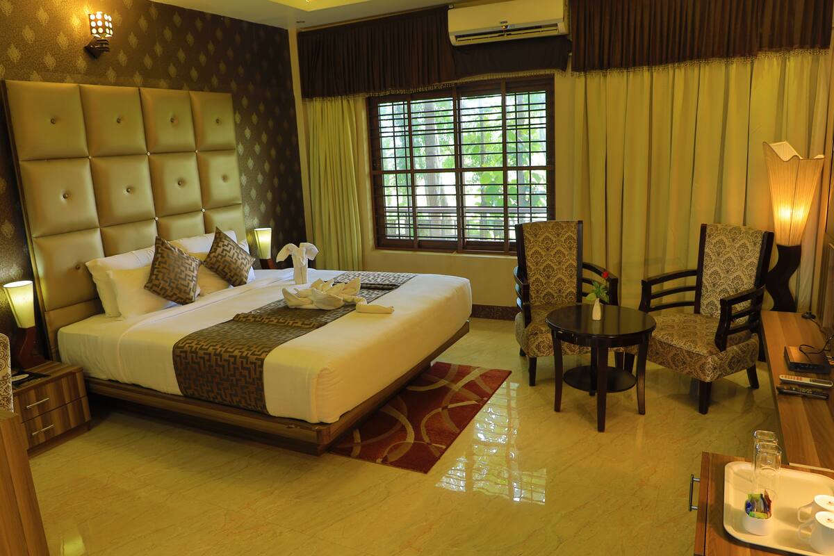 A hotel room with a large bed, a dining table, two chairs, a small table with a tray, and a window with curtains.