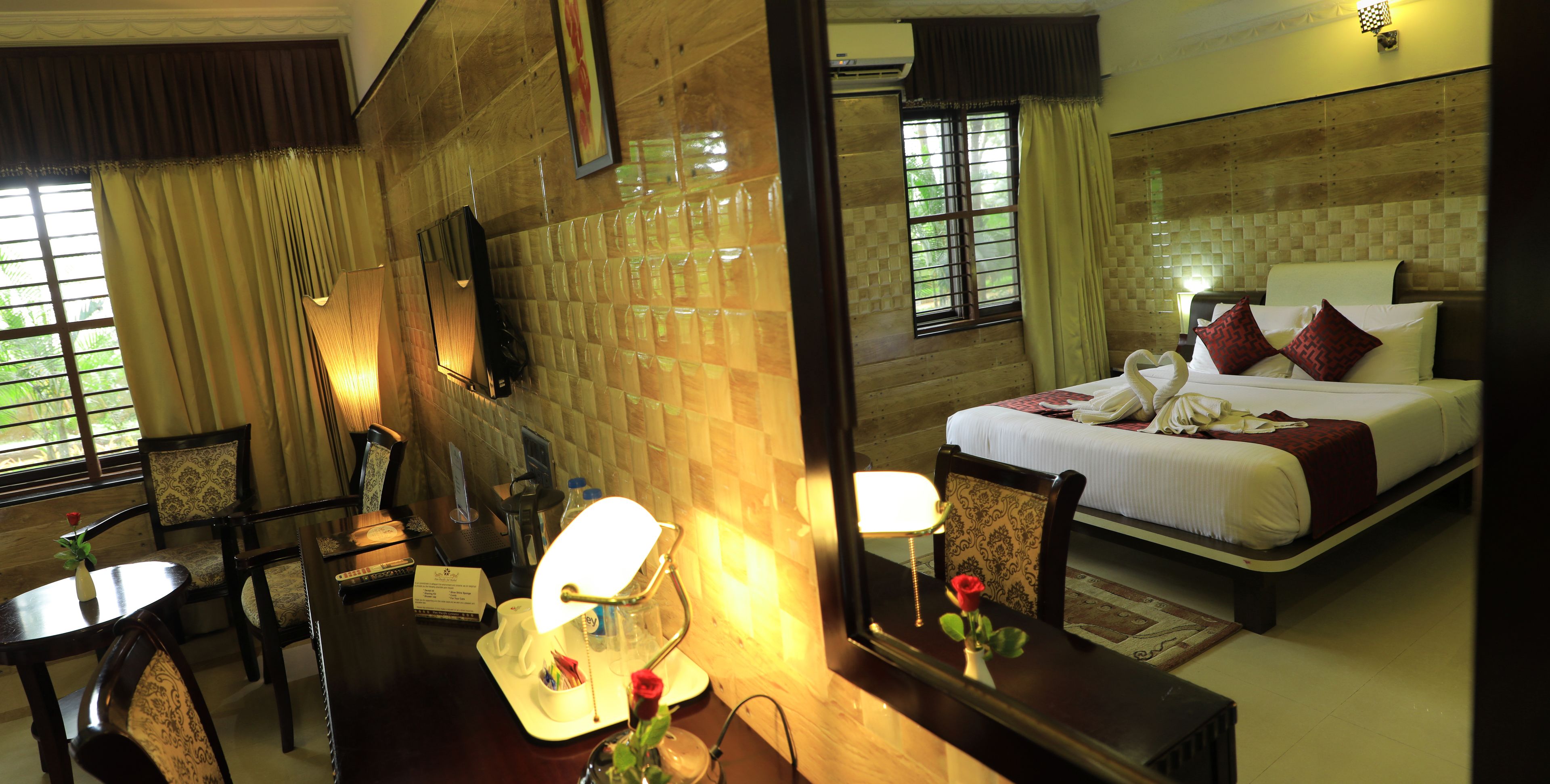 A hotel room with a bed, a desk with a lamp, a chair, a mirror, and a vase with a rose.