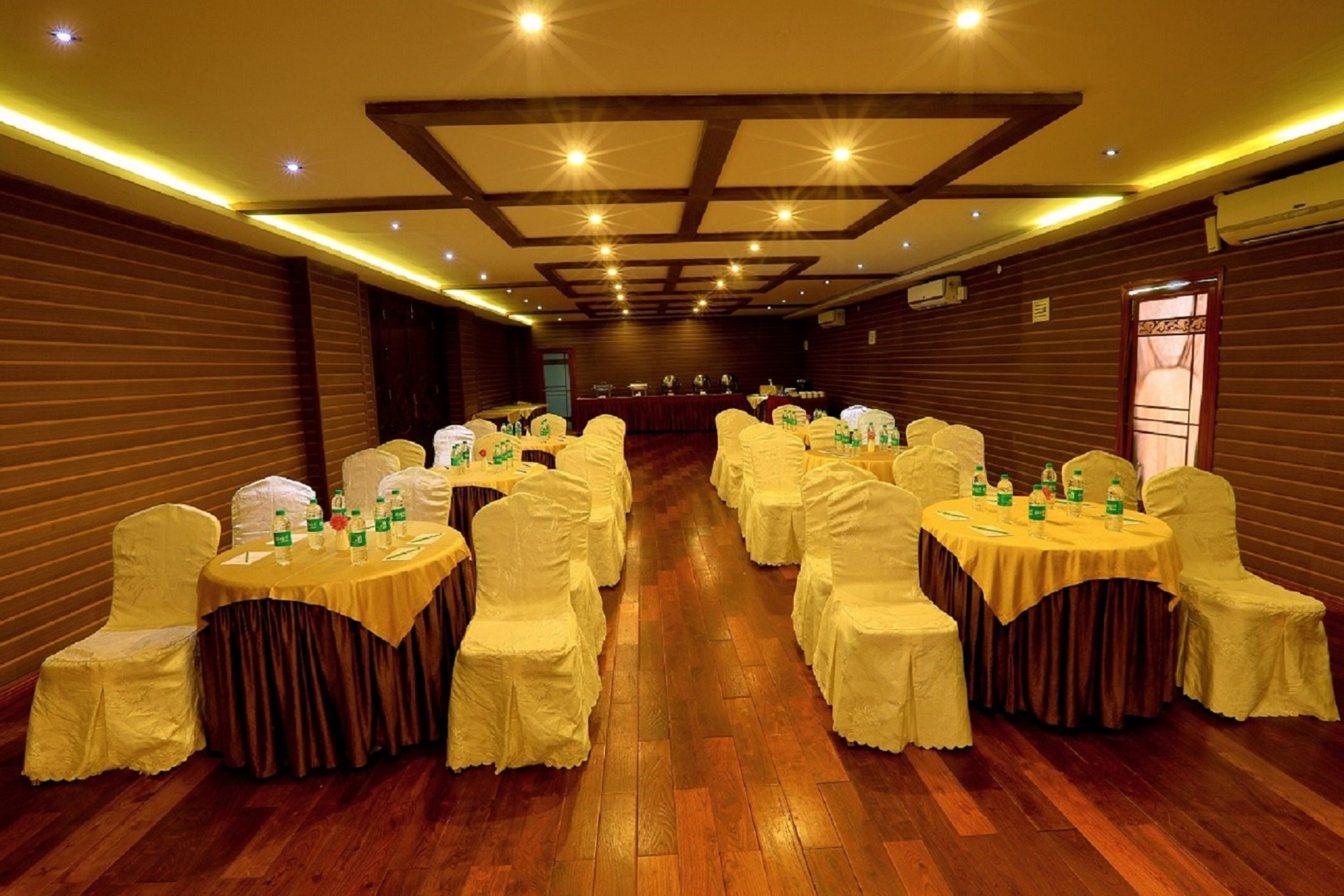 A banquet hall with round tables and chairs, wooden flooring, and a high ceiling with recessed lighting.