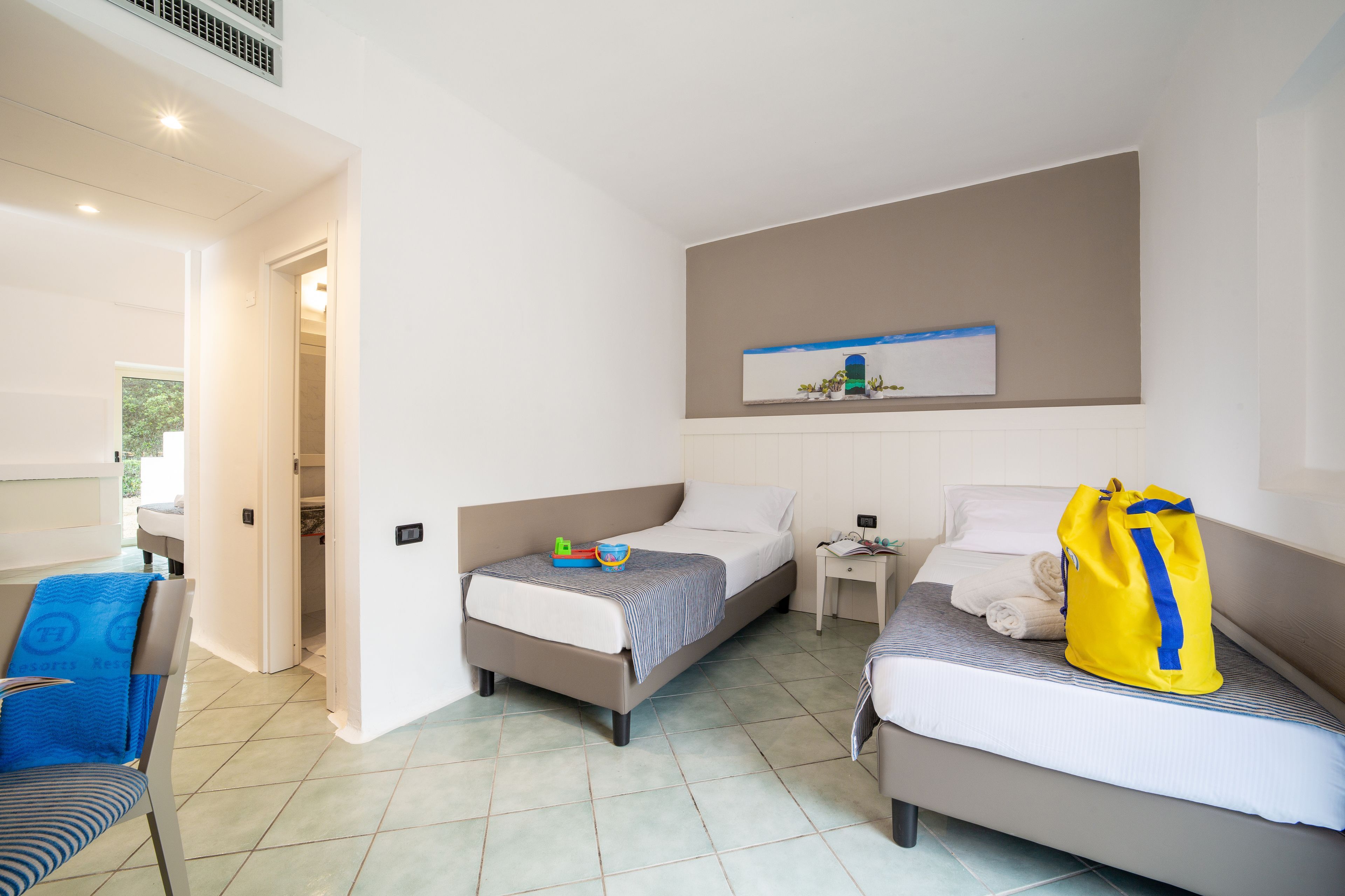 Habitación de hotel con dos camas, una silla, una mesita pequeña y una obra de arte colgada en la pared.