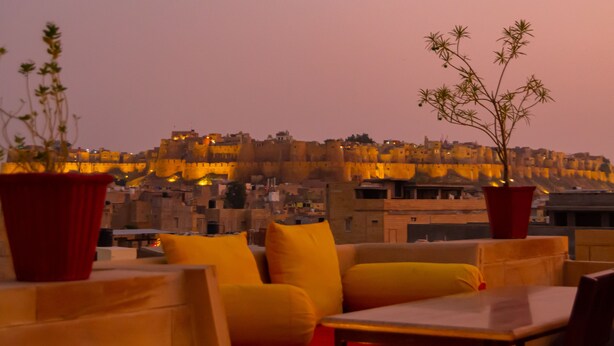 Terraza en la azotea con cojines amarillos y macetas rojas, con vistas a una ciudad amurallada histórica al atardecer.