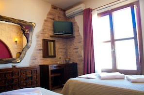 Habitación de hotel con pared de piedra, televisor montado, cómoda de madera, cama con ropa blanca y ventana con cortinas.