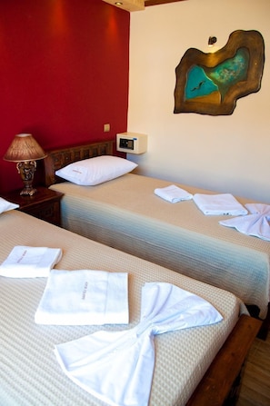 Habitación de hotel con dos camas, cada una con un almohadón blanco y una toalla doblada. Tiene un cabecero de madera, una lámpara sobre una mesita de noche y un cuadro colgado en la pared.