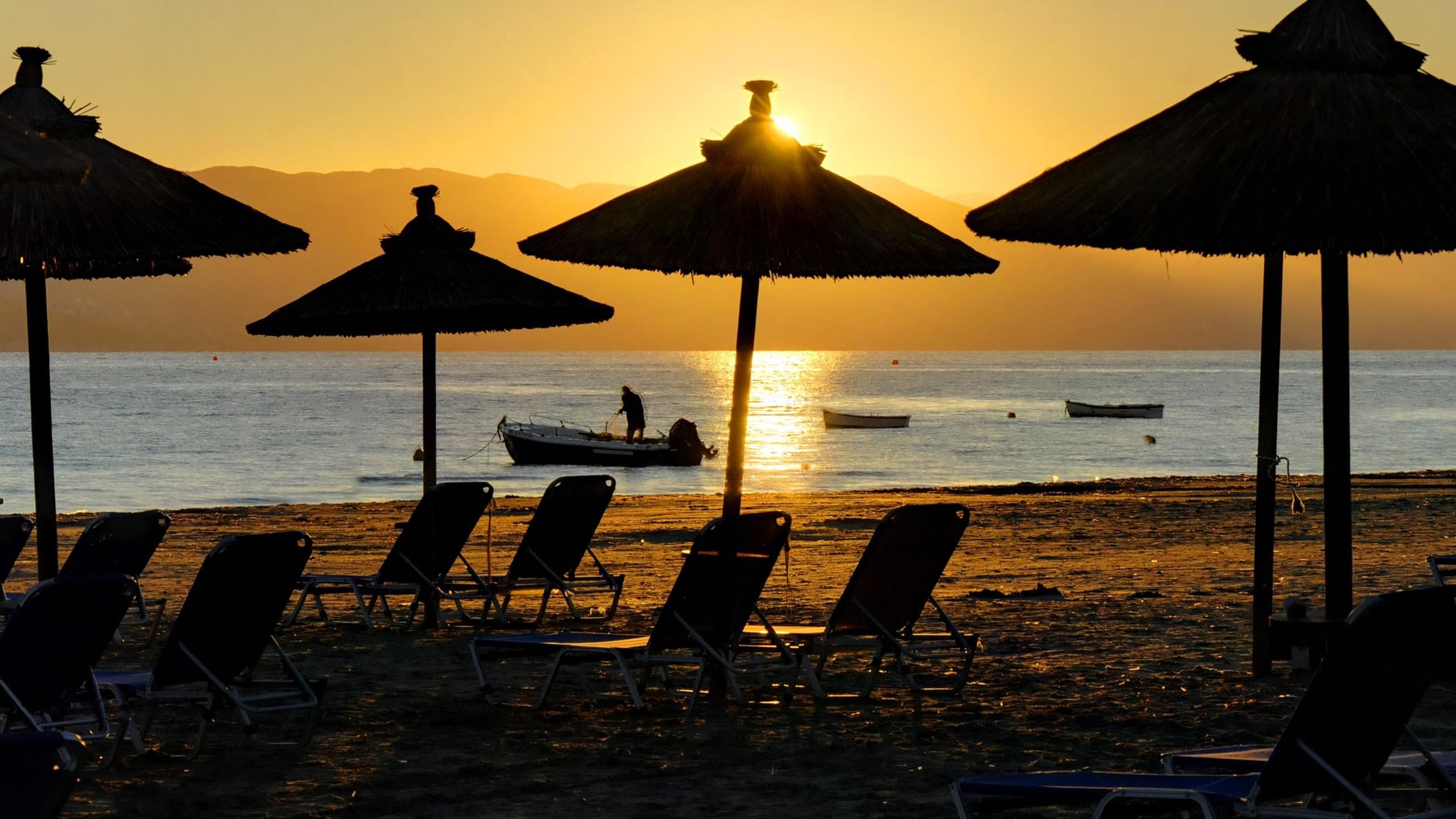 A beach with lounge chairs and thatched umbrellas, a person fishing, and a boat on the water during sunset.