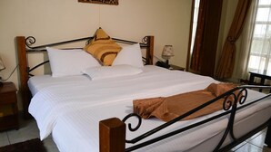 A neatly made bed with white bedding and a brown throw blanket, a wooden headboard, and a small bedside table with a lamp. - Golf Hills Residence Hotel (Kigali)