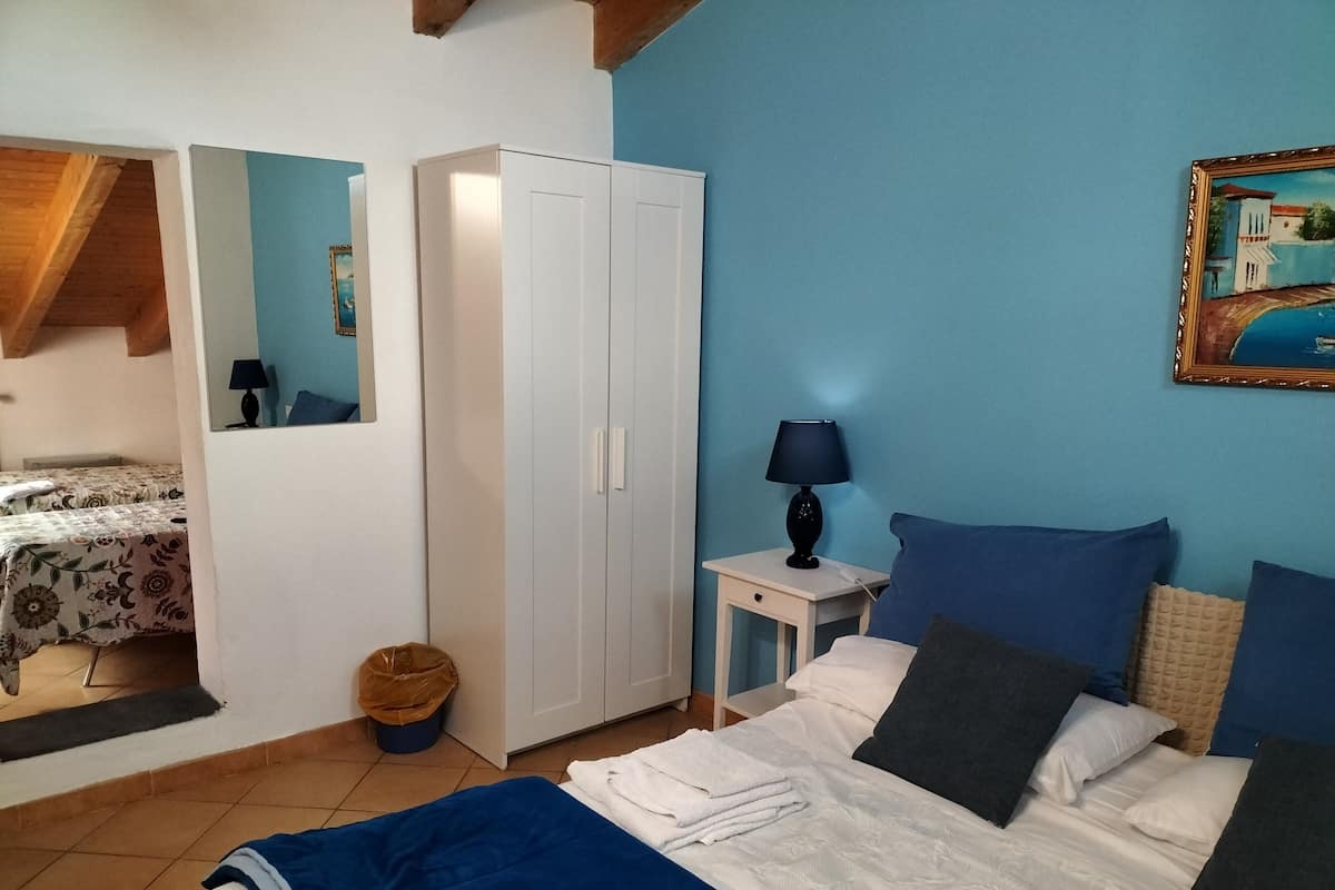 Ein Schlafzimmer mit Bett, Kleiderschrank, Nachttisch mit Lampe, einem Bild an der Wand und einem Fenster mit Blick auf ein Gebäude.