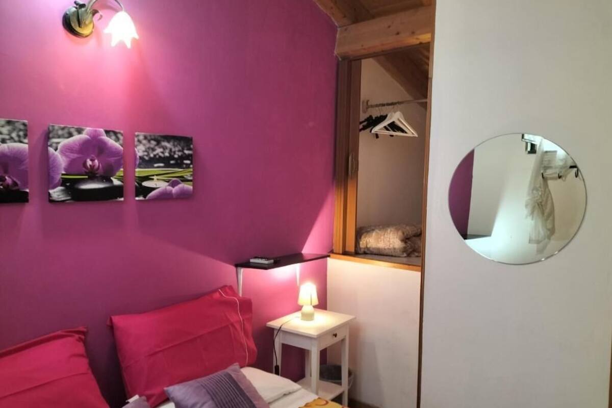 Ein Schlafzimmer mit pinkfarbener Wand, einem Bett mit roten und violetten Kissen, einem Nachttisch mit Lampe, einem Spiegel und einem Fenster mit Holzrahmen.