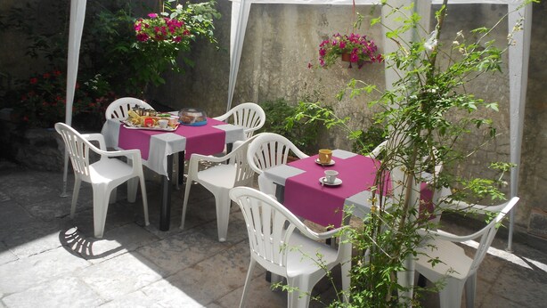 A covered outdoor dining area with white plastic chairs and tables, surrounded by greenery and a stone wall. - Da Rosa (Linguaglossa)