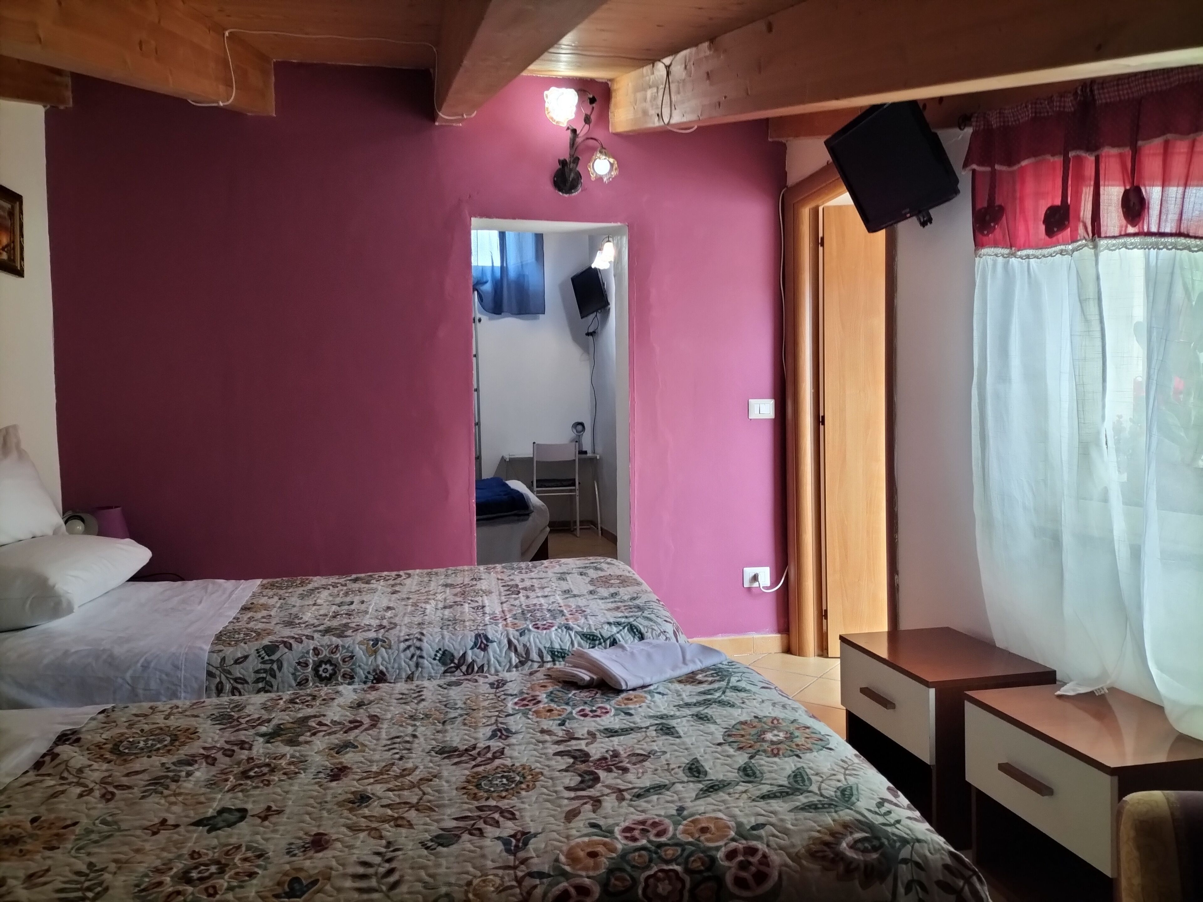 A bedroom with a floral bedspread, a wooden ceiling, a TV on the wall, and a window with sheer curtains.