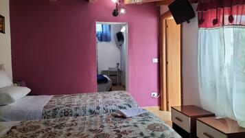 A bedroom with a floral bedspread, a wooden ceiling, a TV on the wall, and a window with sheer curtains.