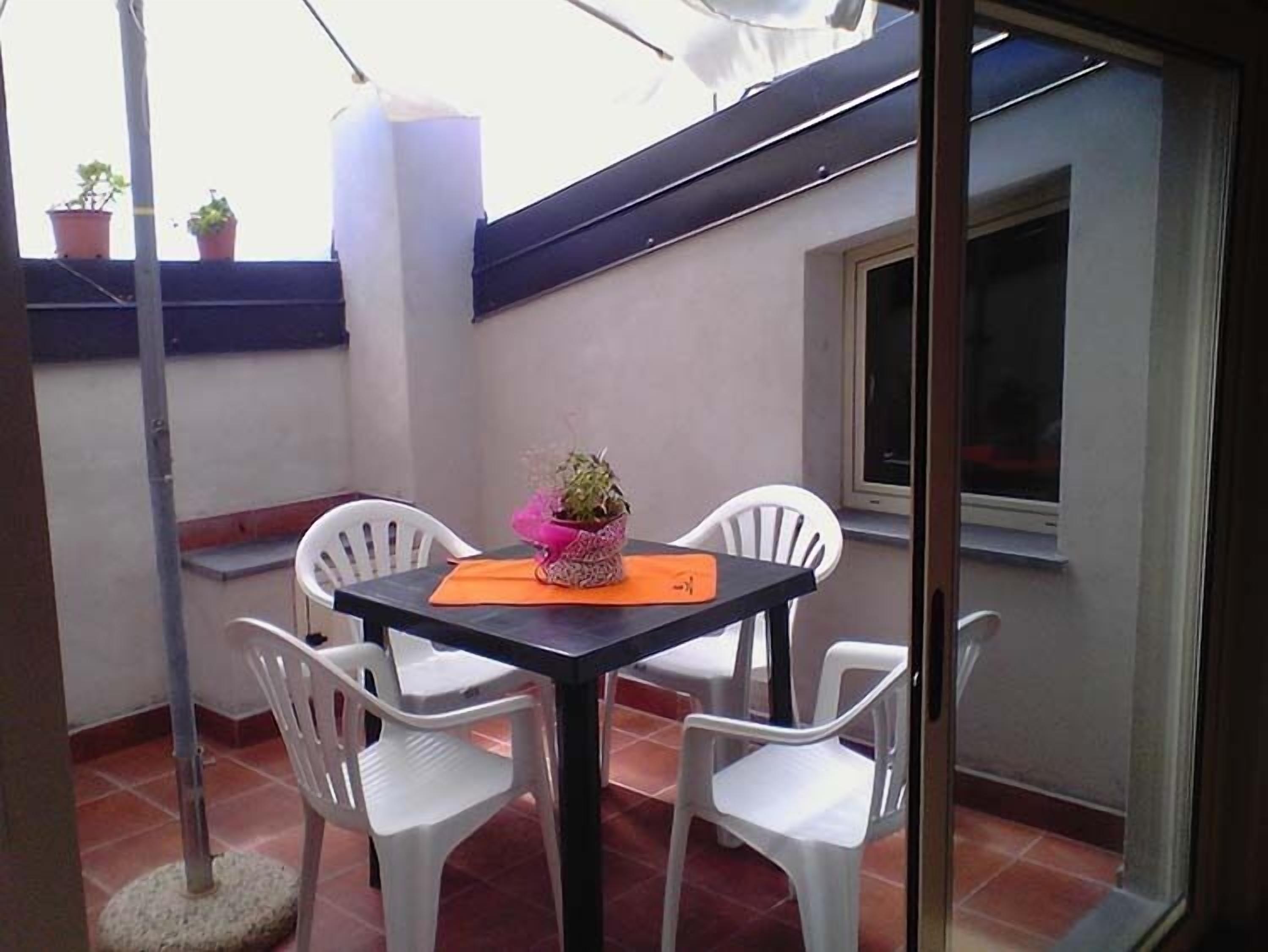 Een klein terras buiten, met een tafel en vier witte stoelen, een bloempot en een raam.