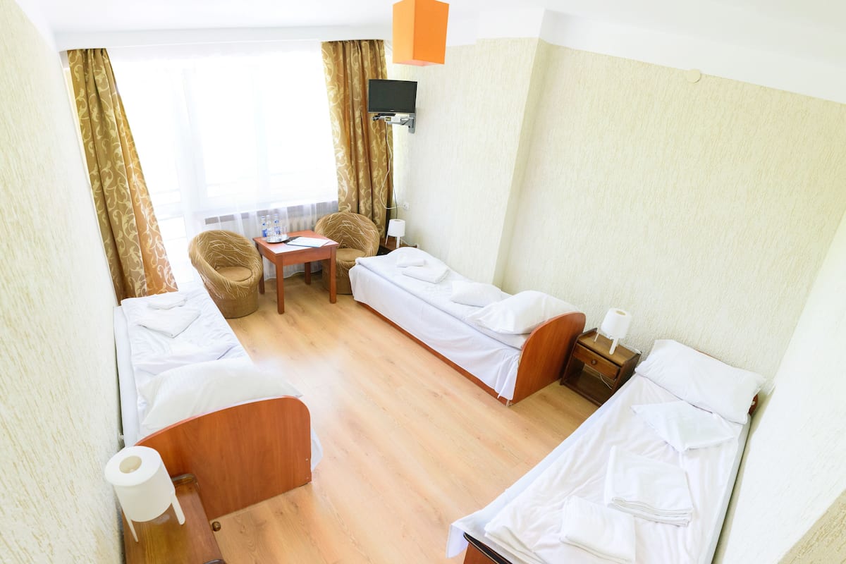 Quarto de hotel com duas camas de solteiro, uma mesa pequena com uma cadeira, televisão fixada na parede e janela com cortinas.