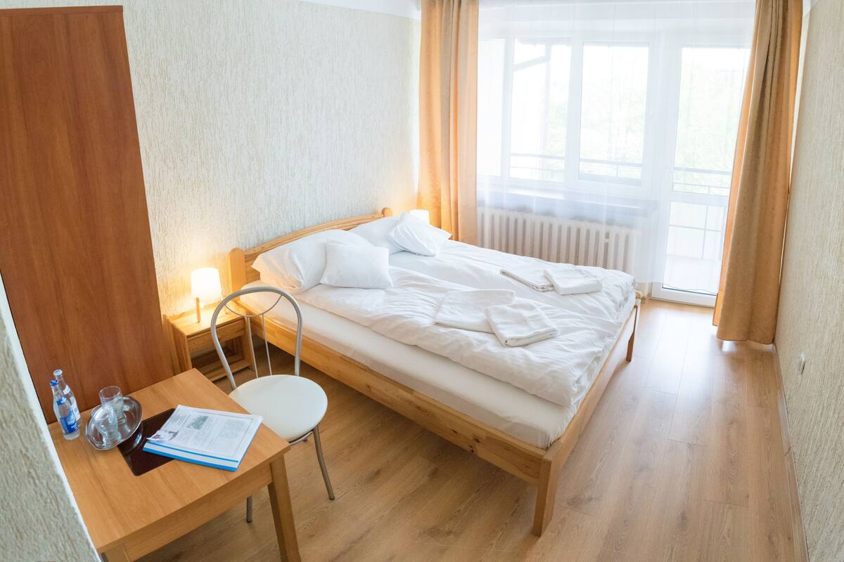 Quarto de hotel com cama de madeira, secretária com cadeira, janela com cortinas e radiador.
