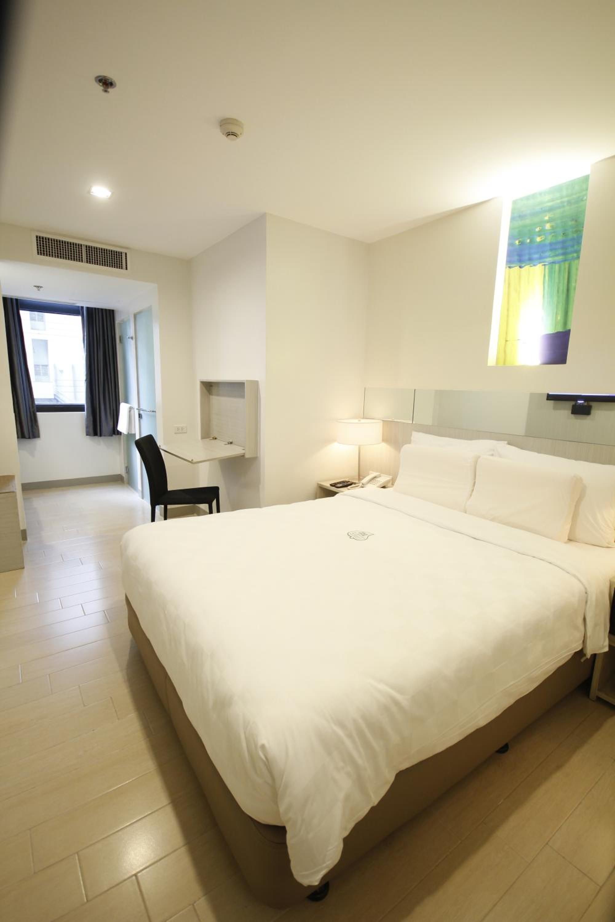 Habitación de hotel con una cama grande, un escritorio, una silla, una mesita de noche con lámpara y una ventana con cortinas.