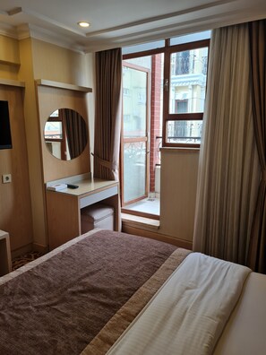 Hotellihuone, jossa on sänky, kirjoituspöytä peilillä, ikkuna verhoilla ja näkymä ulkona oleviin rakennuksiin.