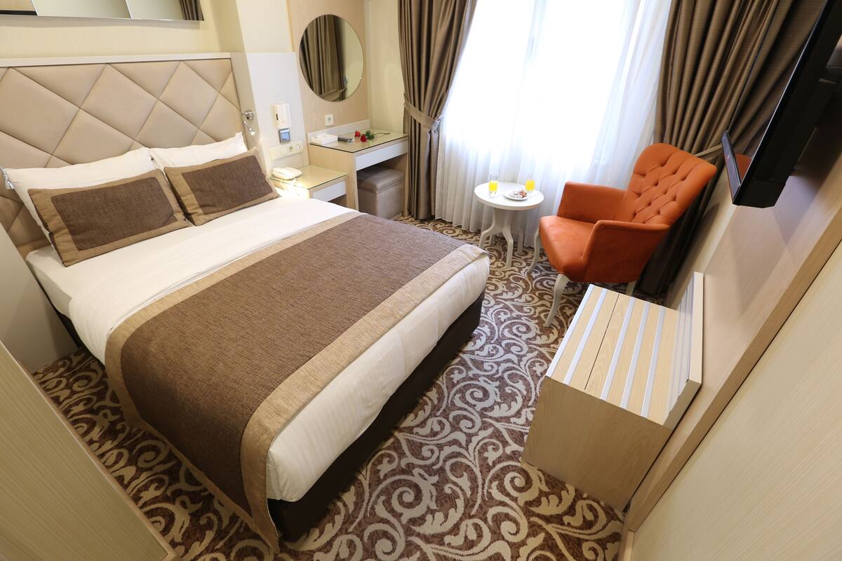 A hotel room with a large bed, a desk, a chair, a TV, and a small table with a glass of orange juice.