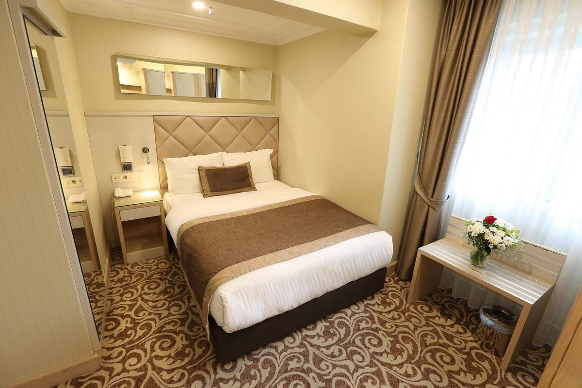 A hotel room with a bed, bedside tables, a mirror, a small table with a vase, and a patterned carpet.