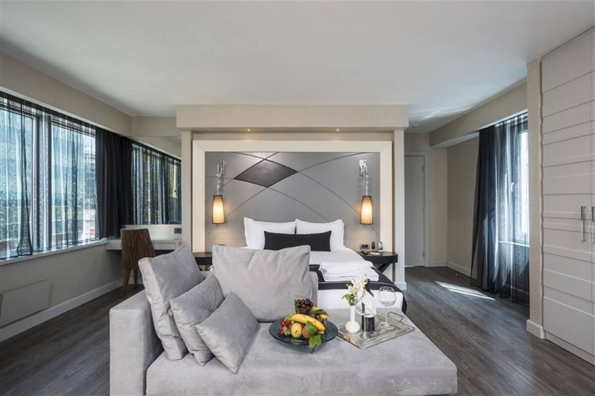 Une chambre d’hôtel moderne avec un grand lit, un canapé gris et une table à manger agrémentée de fruits et d’un verre.