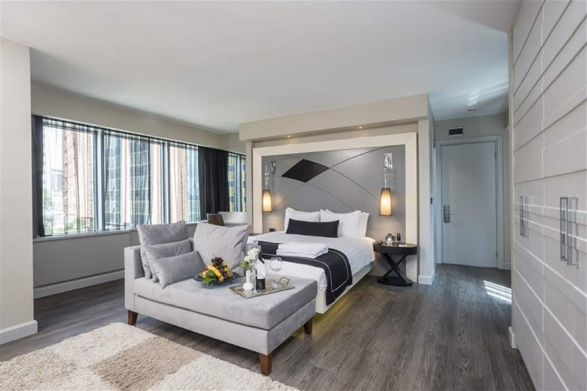 Une chambre d’hôtel moderne avec un grand lit, un canapé et une table à manger. On y trouve une fenêtre avec des rideaux, un luminaire fixé au mur et une porte.