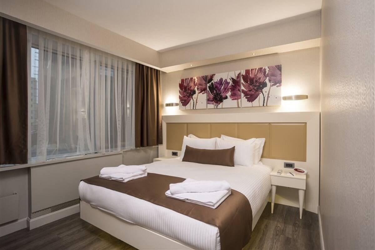 Une chambre d’hôtel avec un lit, une table de chevet, une œuvre d’art accrochée au mur et une fenêtre avec des rideaux.