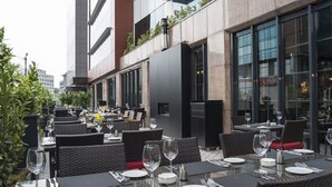 Un espace extérieur aménagé pour les repas, avec des tables et des chaises, un bâtiment moderne aux fenêtres en verre, et une vue sur le paysage urbain.