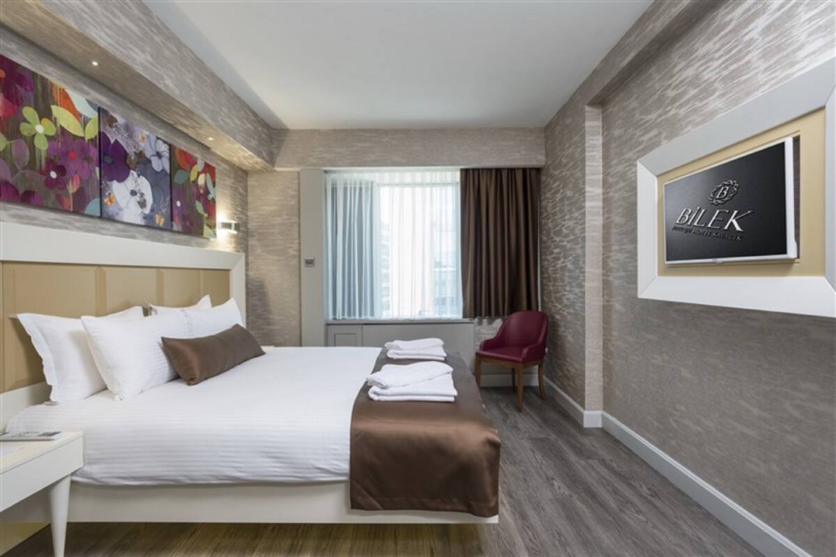 Une chambre d’hôtel moderne avec un grand lit, un fauteuil rouge, une fenêtre avec des rideaux et un panneau fixé au mur.