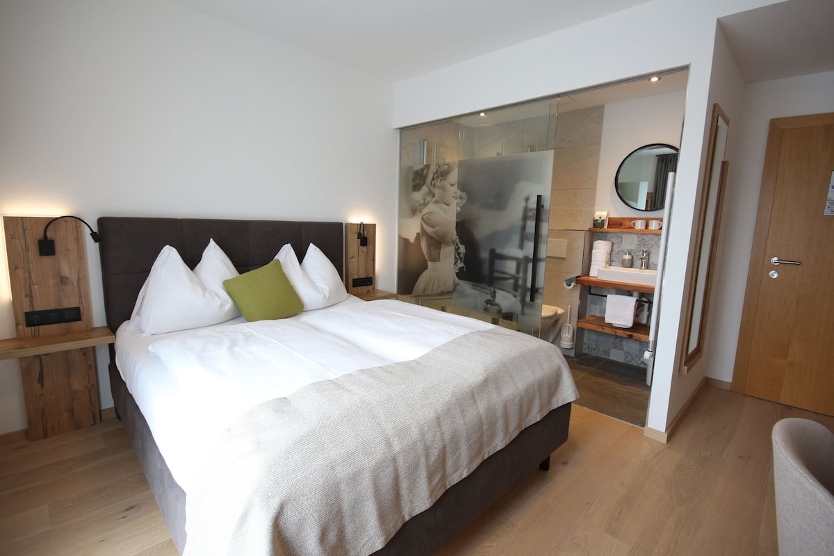 Une chambre d’hôtel moderne dotée d’un grand lit, de tables de chevet, d’une salle de bain avec une douche à parois de verre et d’une porte en bois.