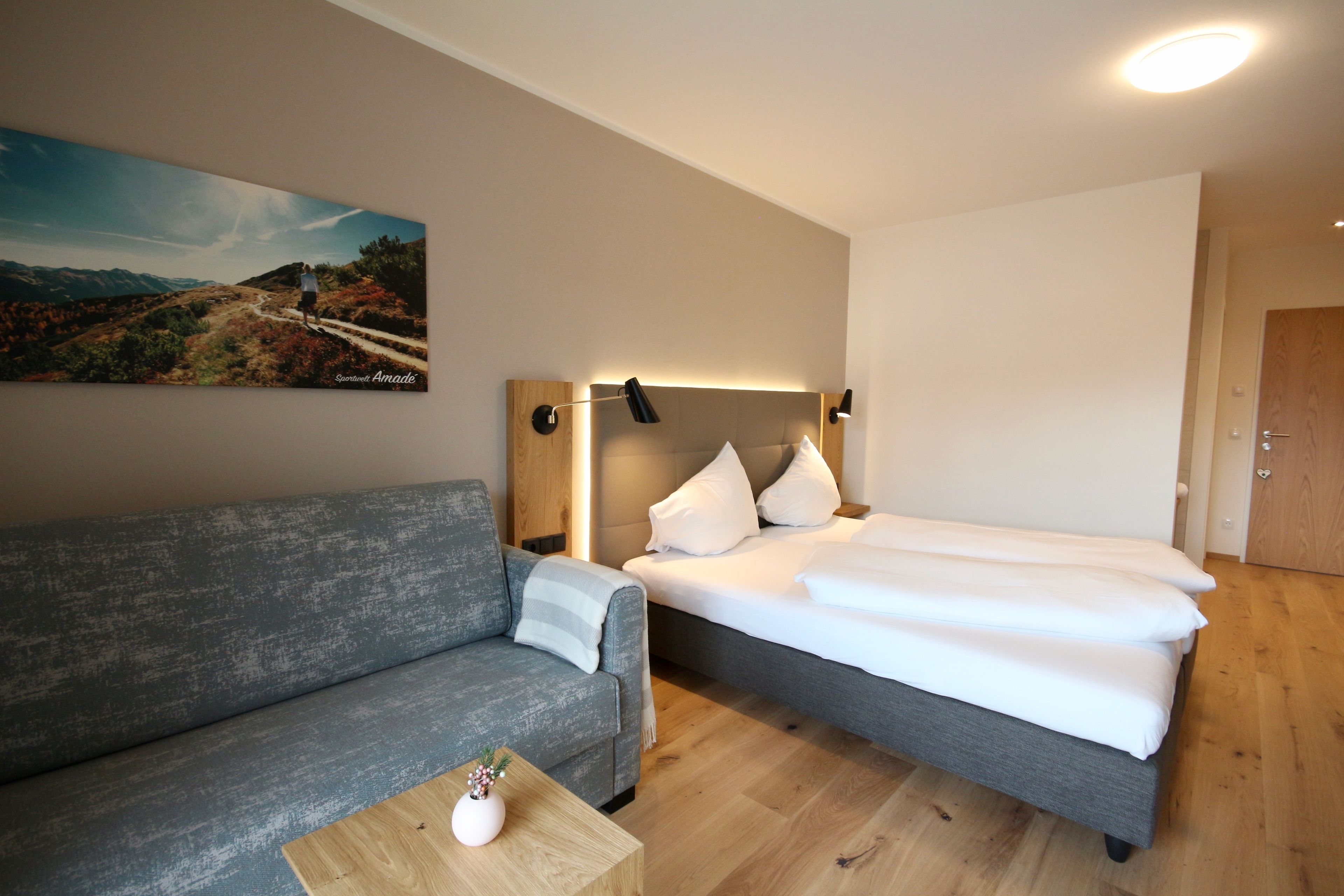 Una habitación de hotel moderna con un sofá gris, una cama con ropa blanca, una mesa de centro de madera con un florero y un cuadro de un paisaje en la pared.