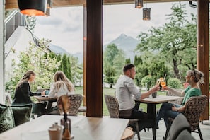 Un café al aire libre con mobiliario de mimbre, vistas a zonas verdes y montañas, y luces colgantes.