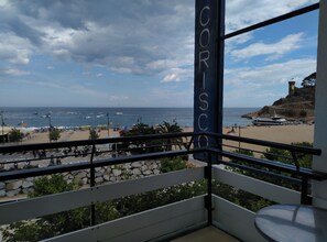 A balcony with a view of the beach, a stone wall, and a flag. - Hotel Corisco (Tossa de Mar)