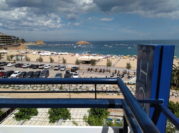 A beachside view with a parking area, beach umbrellas, and boats in the water. - Hotel Corisco (Tossa de Mar)