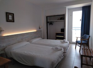 A hotel room with two beds, a chair, a small table, a lamp, and a view of the outside.