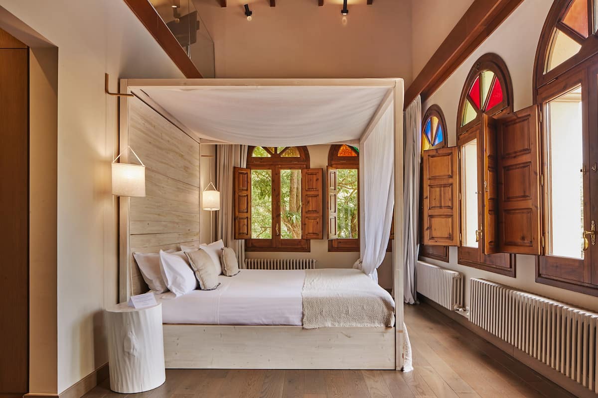 A bedroom with a canopy bed, wooden beams, and a view of greenery through windows.