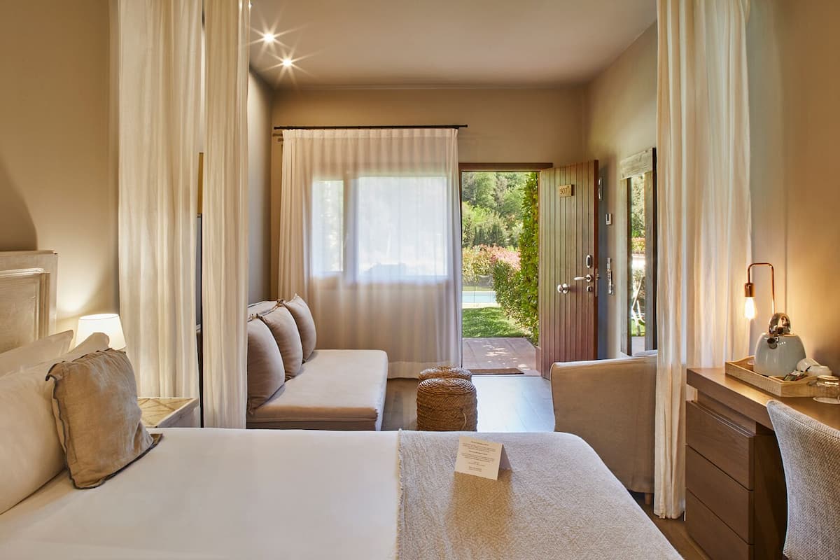 A hotel room with a bed, a sofa, a desk with a lamp, and a view of a garden.