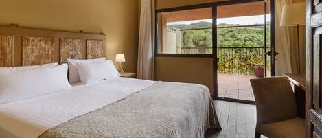 A hotel room with a large bed, a chair, a balcony with a view of greenery, and a window with curtains.