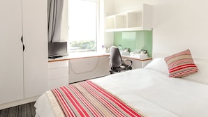 Hotellihuone, jossa on sänky, työpöytä tietokoneella, tuoli ja ikkuna, jossa on verhot.