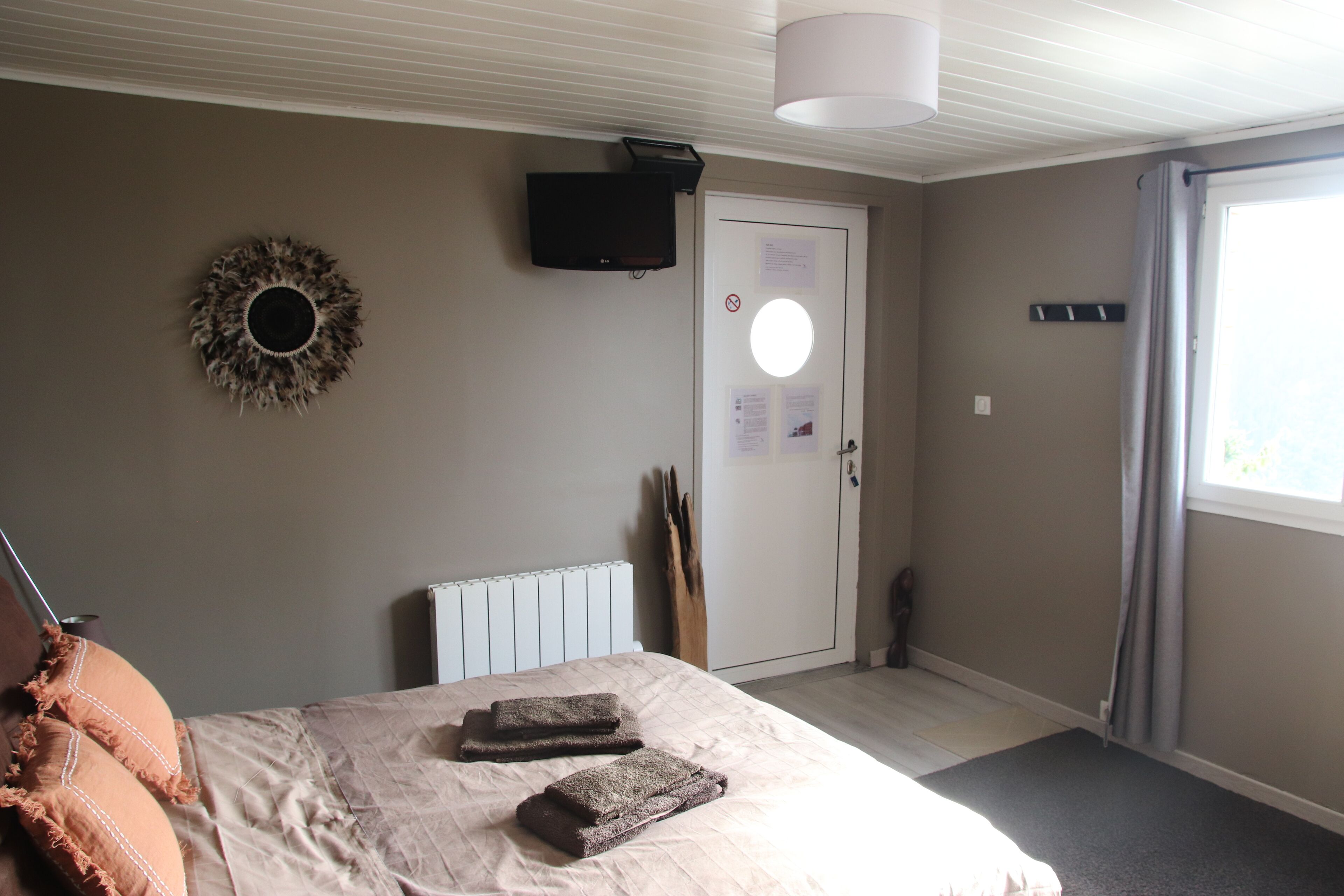 Ett sovrum med en säng, en tv som är monterad på väggen, en dörr med en lapp på och ett fönster med gardiner.