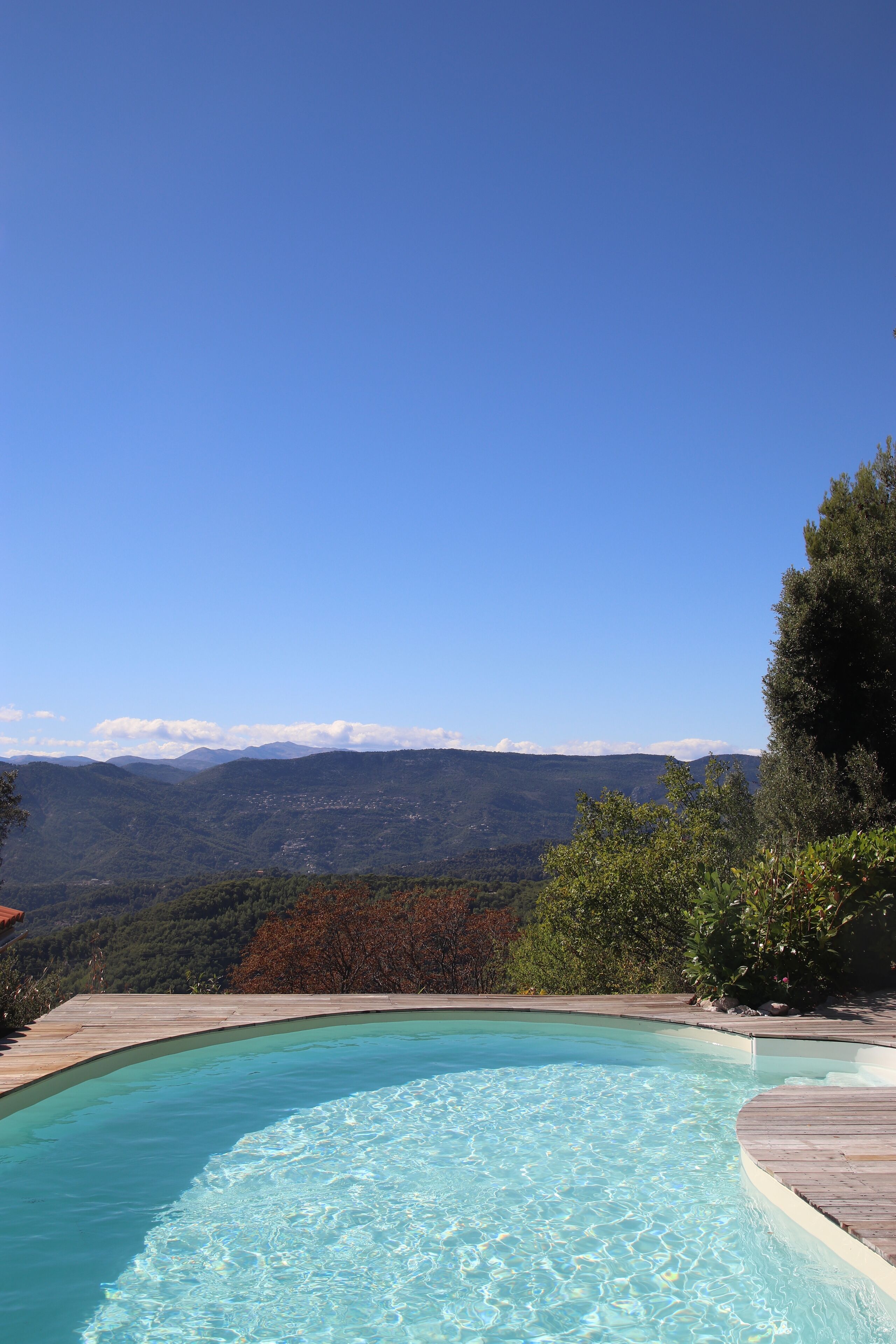 A clear blue sky, a swimming pool with a wooden deck, and a mountainous landscape in the background.