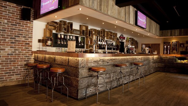 A bar area with a wooden counter, stools, and a menu board. - Becket Court - University of Kent - Campus Accommodation (Canterbury)