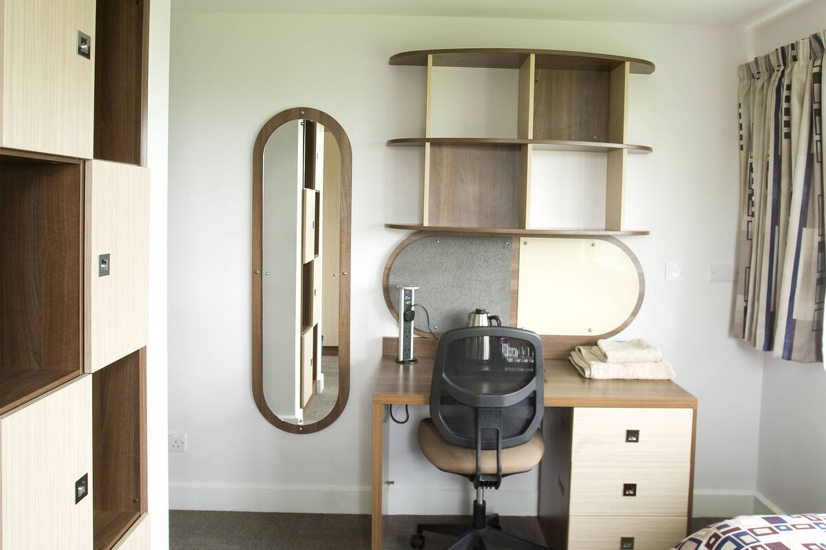 A compact hotel room with a wooden wardrobe, a desk with a chair, a mirror, and a bed with a patterned blanket.