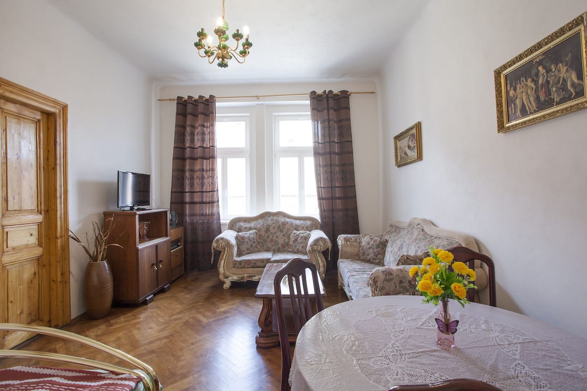 Ein Wohnzimmer mit Holzschrank, Fernseher, zwei Sofas, einem Esstisch mit Blumenvase und einer Chandelier.