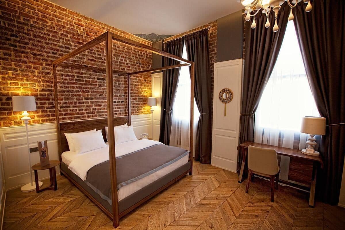 Uma cama de dossel com estrutura de madeira, uma escrivaninha com cadeira e uma janela grande com cortinas.