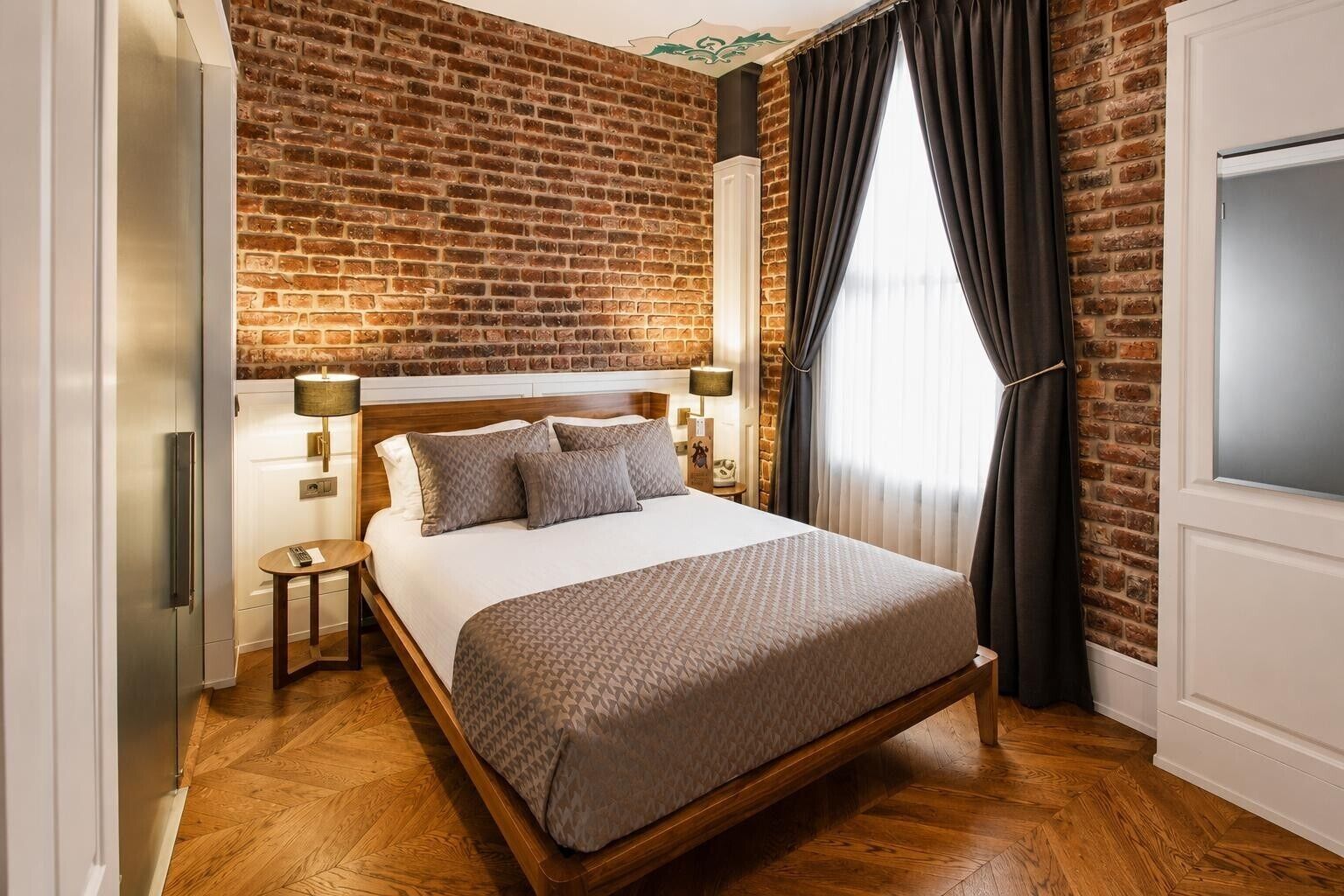 A hotel room with a brick wall, a bed with a wooden headboard, a nightstand, a lamp, and a window with curtains.