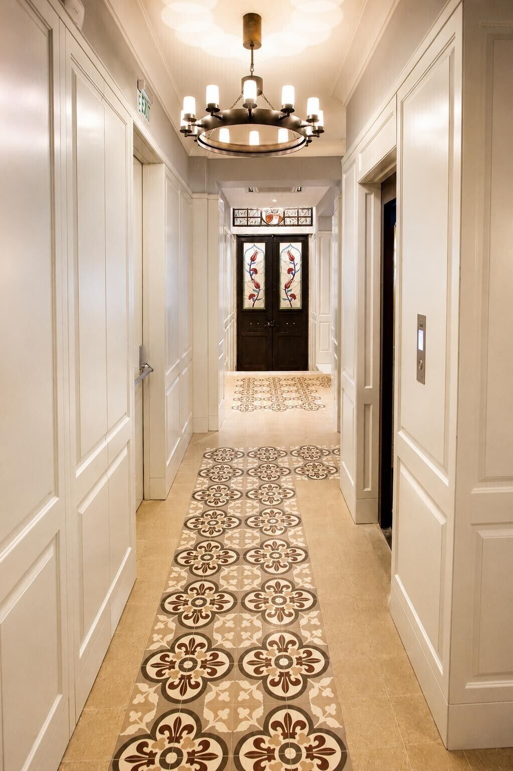 A hotel hallway with a patterned floor, a chandelier, and a double door with a glass panel.