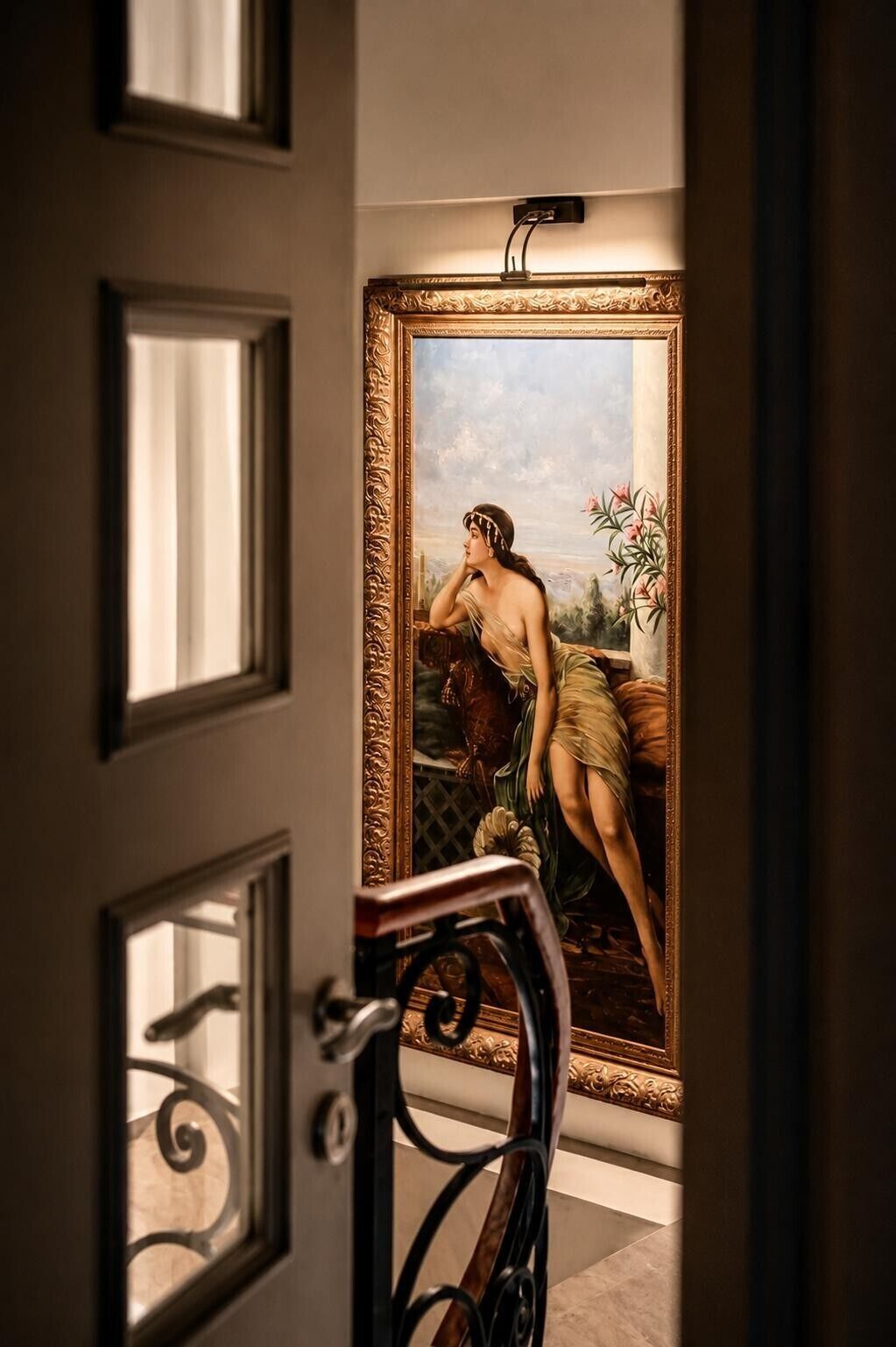 Una habitación con un espejo grande y ornamentado que refleja un cuadro clásico, una puerta con picaporte decorativo, y un vistazo a una escalera.