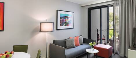 Una habitación de hotel moderna con un sofá gris, una mesita redonda pequeña, una lámpara de pie y un cuadro en la pared.