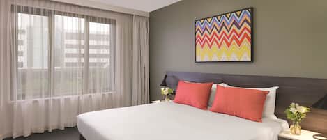 Habitación de hotel con una cama grande, una mesita de noche, una lámpara, una ventana con cortinas translúcidas y un cuadro abstracto de colores en la pared.